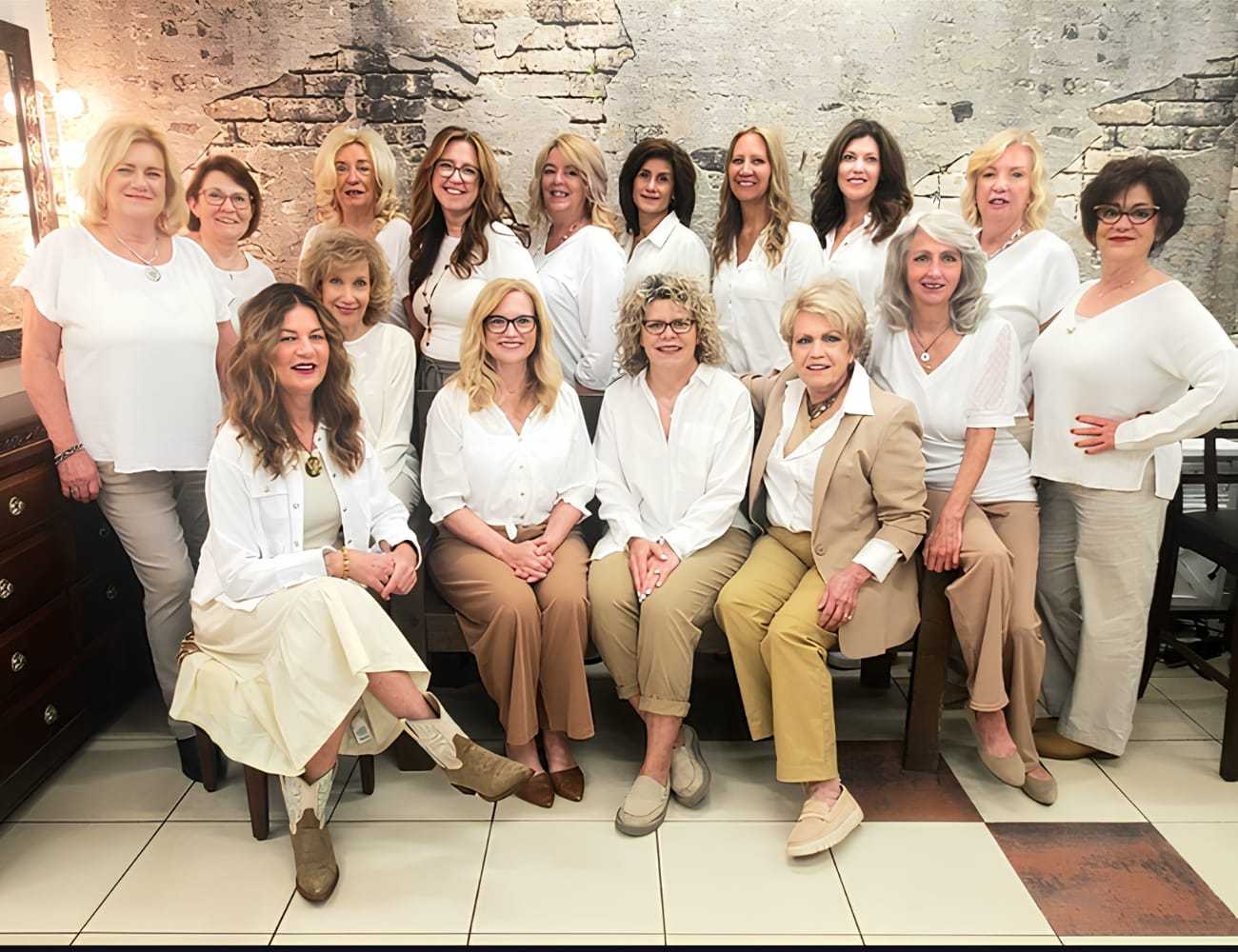 Group of smiling women in coordinated white and beige outfits, posing against a brick wall.
