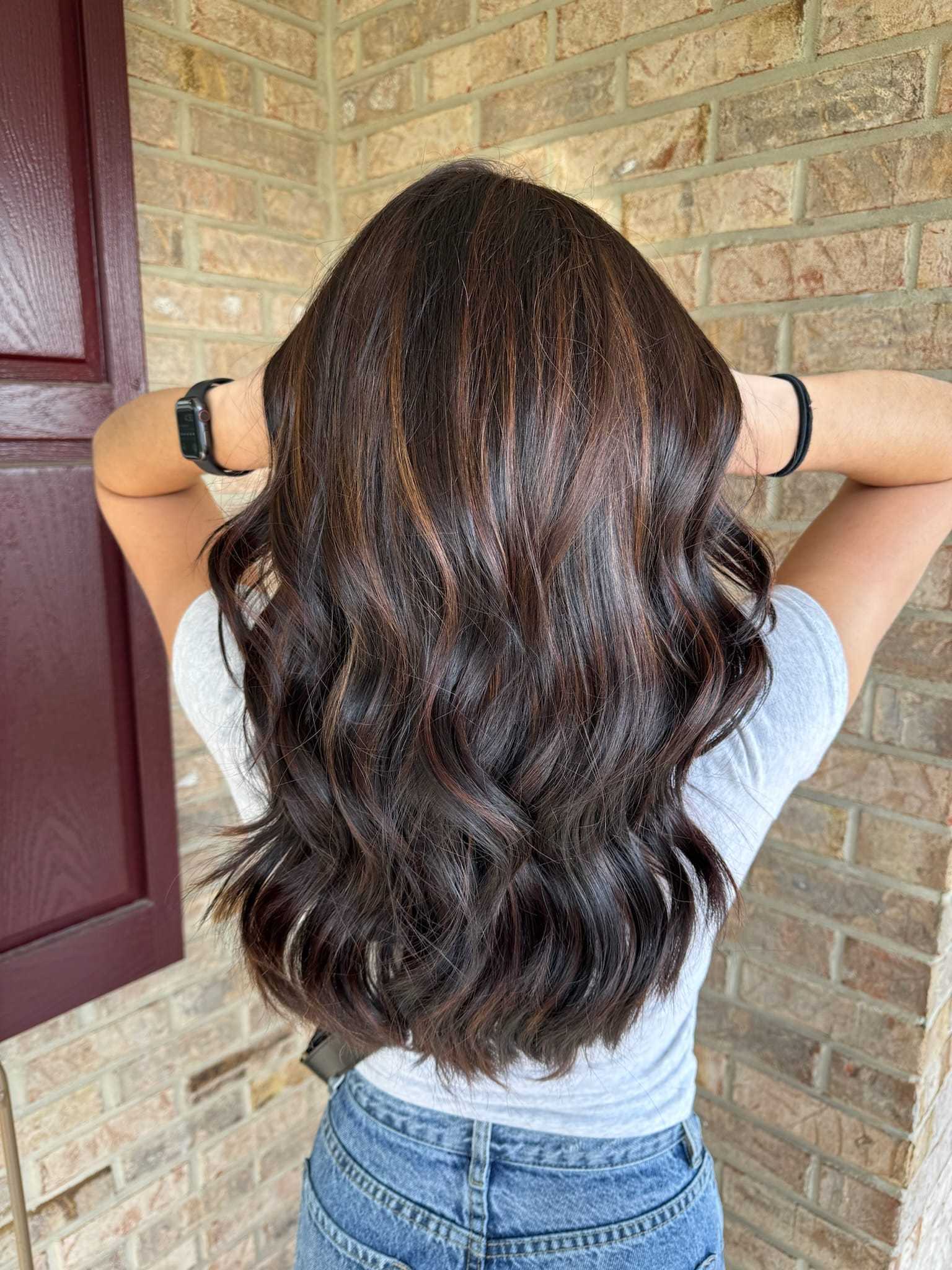 Woman with long, wavy dark hair standing against a brick wall, wearing a gray shirt and jeans.