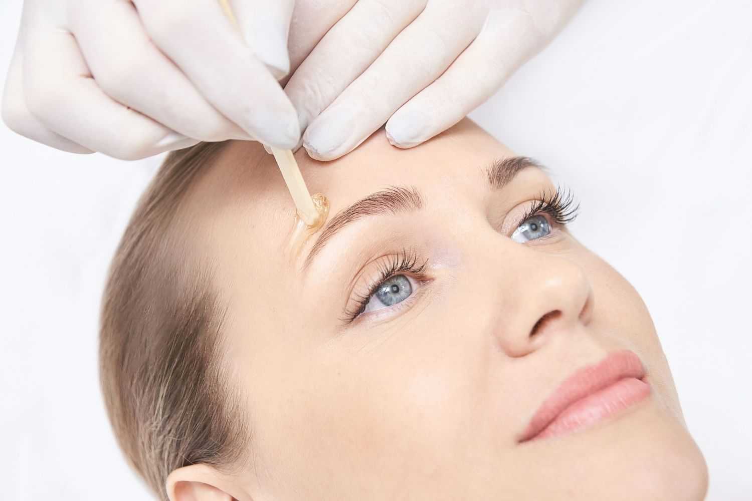 Woman undergoing eyebrow waxing procedure by a professional with a wooden stick.